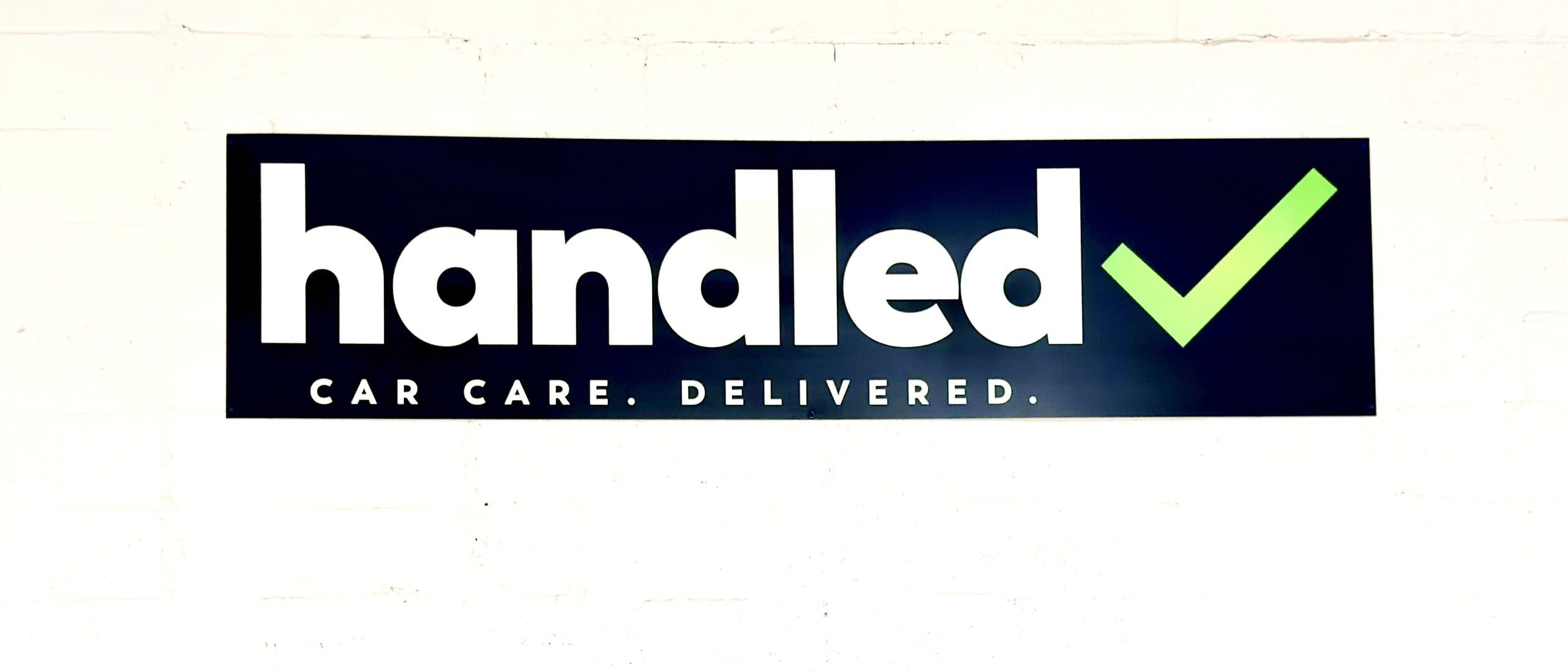 The Handled sign — car care, delivered — on a white block wall
